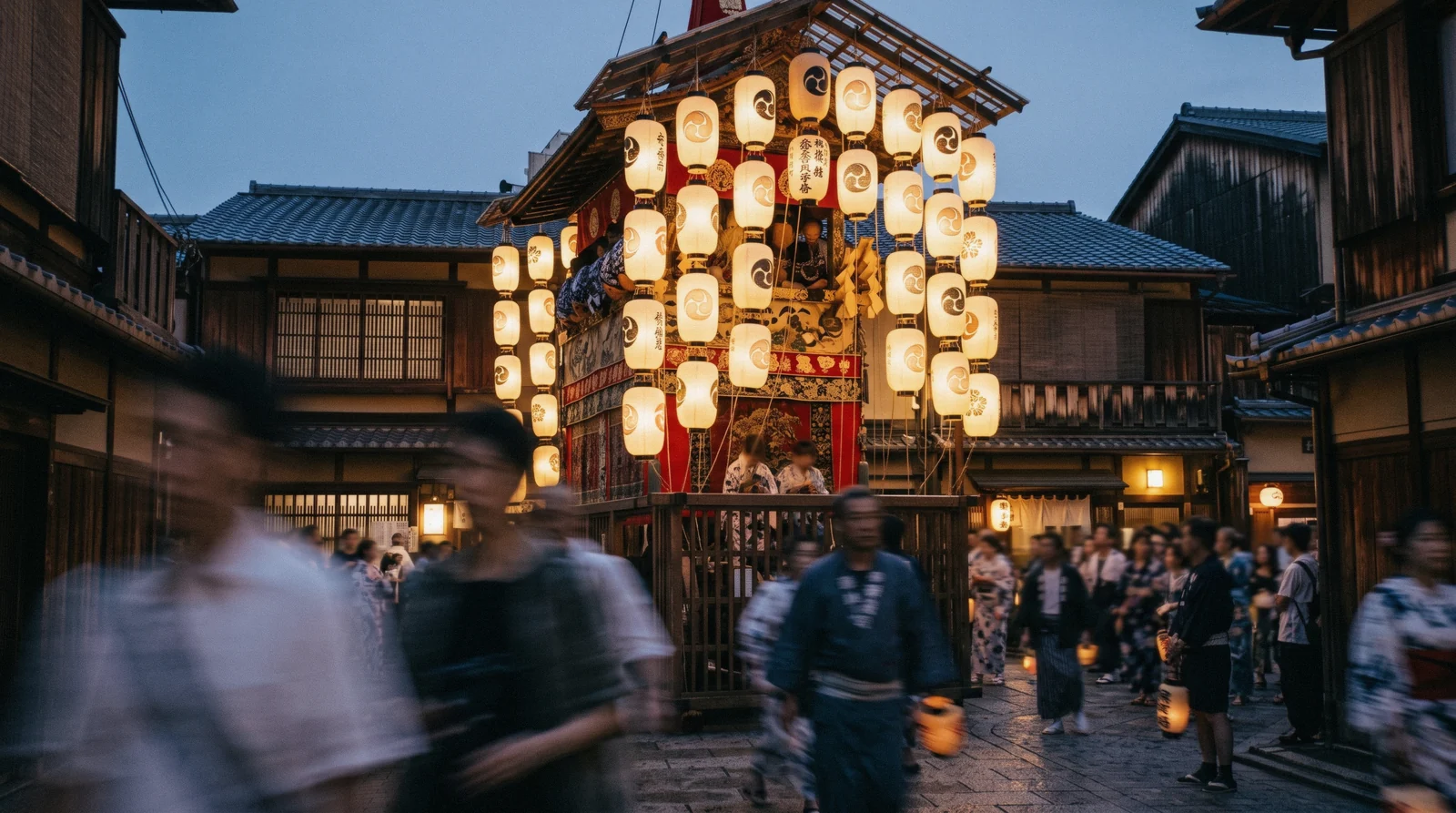 A tall wooden festival float with lanterns being pulled through a city street at dusk, Kyoto festivals Japan